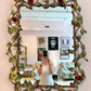 Decorative mirror with floral frame in a room with colorful armchair and framed pictures.